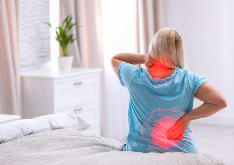 Woman sitting on bed with neck and lower back pain highlighted in red
